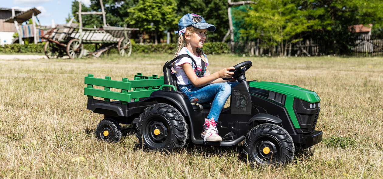 Mädchen fährt mit dem Elektro-Kindertraktor und Anhänger über eine Wiese